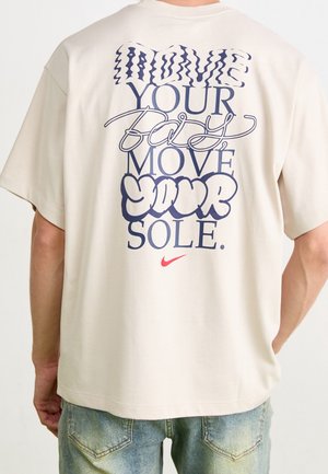 T-shirt blanc avec texte bleu « Move your body move your sole » en polices variées et un petit logo rouge Nike, porté par une personne en jean bleu.