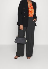 Blazer negro con botones dorados, blusa impresa naranja vibrante, pantalones grises sueltos y un bolso estructurado negro con un asa corta.