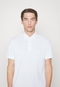 Weißes Poloshirt aus leichtem Baumwollstoff, mit einem knöpfbaren Kragen, kurzen Ärmeln und einem dezenten Logo auf der Brust.