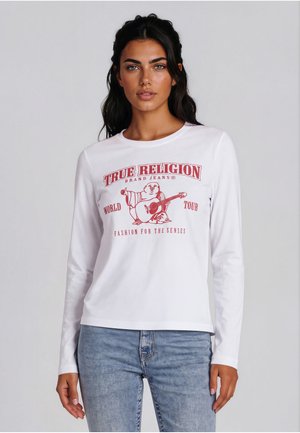 Femme aux cheveux foncés portant une chemise blanche à manches longues avec le logo rouge True Religion et un jean bleu clair sur un fond uni.
