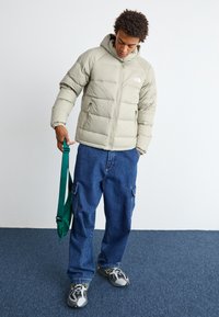 Svijetlosiva puffer jakna s kapuljačom i logotipom, uparena s ležernim plavim cargo trapericama. Model drži zeleni ranac u jednoj ruci.