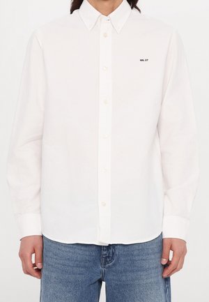 Chemise blanche à manches longues boutonnée avec col, comportant un petit texte "NN. 07" sur la poitrine gauche, portée avec un jean bleu clair.
