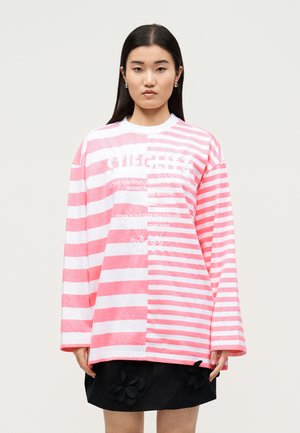 Junge Frau trägt ein übergroßes pink-weiß gestreiftes Langarmshirt mit Schriftzug und einen schwarzen Rock mit Blumenapplikationen.