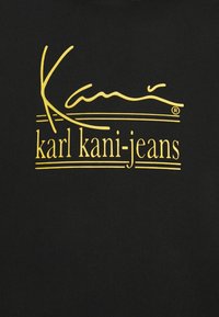 Musta T-paita, jossa on näkyvä keltainen logomuotoilu, joka sisältää kaunokirjoitustekstiä ja brändin nimen "karl kani-jeans." Puuvillakangas.