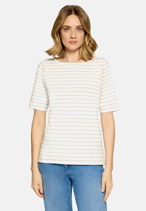 Kvinde med skulderlangt blondt hår iført en hvid kortærmet skjorte med beige striber og blå jeans, stående mod en ensfarvet baggrund.