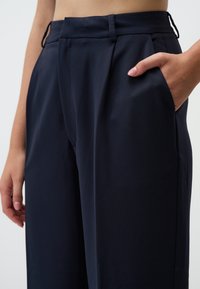 Pantalon taille haute bleu marine avec une texture lisse, devant plissé, poches latérales et passants de ceinture. Main reposant dans la poche.