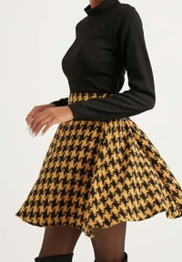 Mujer vistiendo una blusa negra de manga larga metida dentro de una falda acampanada con patrón de damas de honor en amarillo y negro, con medias negras y botas.