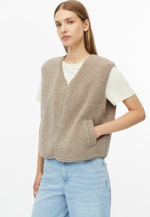 Frau mit langen Haaren, die eine beige strukturierte Weste über einem weißen kurzärmeligen Hemd und hellblauen Jeans trägt, steht mit einer Hand in der Tasche.