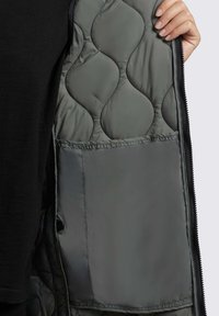 Geraffte graue Steppjacke mit glatter Innenseite. Verfügt über eine große Tasche und sichtbare Nähte entlang der Nähte.