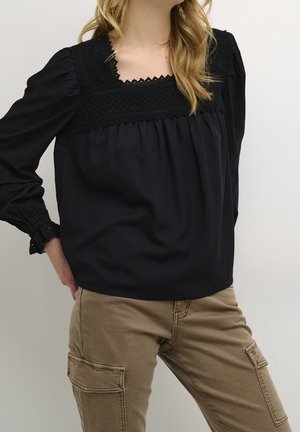 Mujer con blusa negra de manga larga con detalles de encaje combinada con pantalones cargo beige, de pie con una mano en la cadera contra un fondo liso.