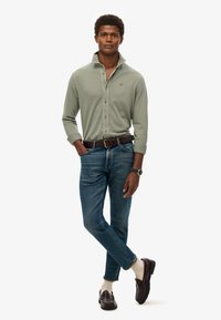 Helles grünes Langarmhemd mit Kragen, kombiniert mit blauen Slim-Fit-Jeans und dunkelbraunen Slippern; beinhaltet einen braunen Gürtel und eine silberne Uhr.