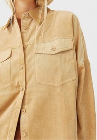 Tan corduroy shirt met een knoopsluiting aan de voorkant, twee borstzakken en lange mouwen. De stof heeft een gestructureerd, ribbelontwerp.