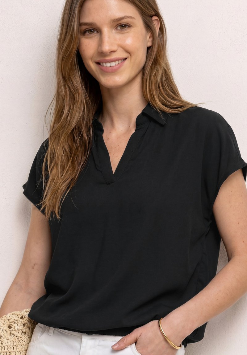 Jeune femme aux longs cheveux châtain clair portant un chemisier noir à manches courtes, un pantalon blanc et un bracelet en or, souriant devant un mur clair.