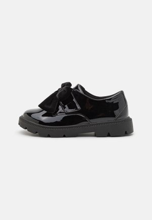 Chaussure en cuir verni noire avec semelle épaisse en caoutchouc et grand nœud en velours noir sur le dessus, montrée de côté sur fond blanc.