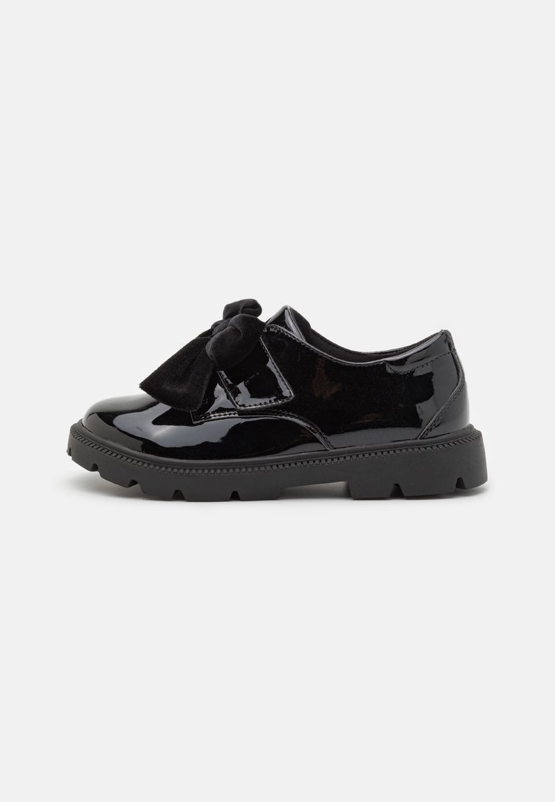 Chaussure en cuir verni noire avec semelle épaisse en caoutchouc et grand nœud en velours noir sur le dessus, montrée de côté sur fond blanc.
