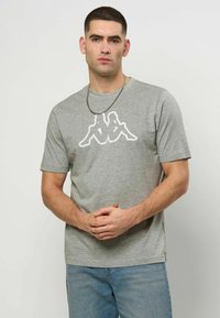 Mann med kort hår iført en grå t-skjorte med en hvit logo av to sittende figurer rygg mot rygg og lys blå jeans, stående med hendene sammenflettet.