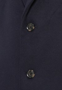 Nahaufnahme eines marineblauen Wollmantels mit zwei schwarzen Knöpfen und einer strukturierten Oberfläche. Der Stoff wirkt weich und leicht flauschig, was Wärme betont.