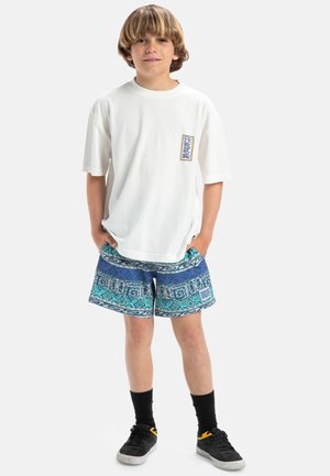 Bílý oversized tričko s grafickou nášivkou, v kombinaci s modrými vzorovanými plavkovými šortkami s geometrickými vzory, nošené s černými ponožkami a teniskami.
