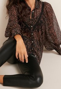 Blouse marron transparent à motifs avec des accents noirs, présentant un devant boutonné, des manches bouffantes et un design texturé. Associée à des leggings en cuir noir.