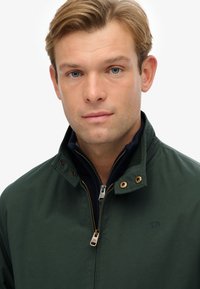 Donkergroene jack met hoge kraag, voorzien van een ritssluiting aan de voorkant, gouden hardware en een klein logo op de borst. Glad textuur.