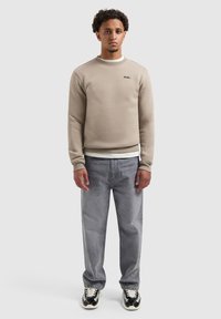 Jonge man in een beige sweatshirt, grijze spijkerbroek en zwart-witte sneakers, staand tegen een plain lichtgrijze achtergrond.