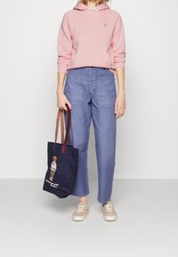 Sudadera de algodón rosa, pantalones de pierna ancha azul claro, zapatillas beige y una bolsa de mano azul marino con un diseño bordado de un oso y asas marrones.