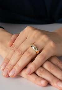 Guldmetallring, slät och rund, bärs på en hand med naturliga, opolerade naglar. Händerna är placerade ihop mot en ljus yta.