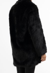 Černý kabát z umělé kožešiny s měkkým, strukturovaným povrchem. Má volný střih, dlouhé rukávy a délku po střed stehen.