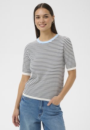 Smilende kvinde med langt mørkt hår iført sølvfarvede øreringe i hoops, en hvid og marineblå stribet kortærmet sweater og blå jeans med hånden i lommen.