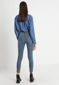 High-waisted blue denim jeans with a slim fit, featuring back pockets and a leather logo patch, paired with a loose blue chambray shirt.