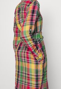 Die Hand einer Frau in einer Tasche, die ein langärmliges, mit einem Gurt versehenes, mehrfarbiges Karokleid mit Druckknopfdetails an den Ärmeln und Manschetten trägt.