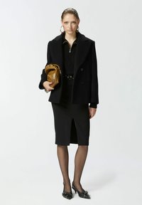 Crni vuneni kaput s izrezanom ovratnikom, uparen s crnim rebrastim džemperom, suknjom olovka i crnim cipelama s šiljastim vrhom. Drži smeđi clutch.