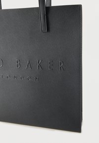 Μαύρη τσάντα ώμου από υλικό με υφή, με ανάγλυφα γράμματα, δομημένο σχήμα και διπλές λαβές για κράτημα.