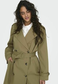 En khakifärgad trenchcoat med dubbelknäppt front, stora knappar, bälte och sidofickor. Lager på en vit skjorta med krage.