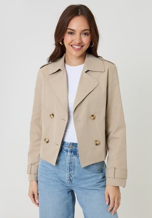 Kvinde smilende iført en beige dobbeltbrystet cropped jakke, hvid top, lyseblå jeans og guldringøreringe mod en ensfarvet baggrund.