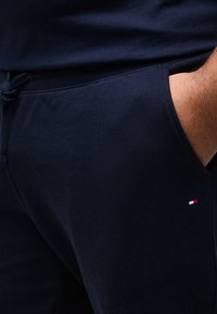 Κοντινό πλάνο χεριού ατόμου στην τσέπη σκούρου μπλε φόρμας με μικρό κόκκινο, λευκό και μπλε λογότυπο κοντά στην τσέπη.