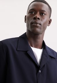 Granatowa kurtka z kołnierzykiem, wykonana z gładkiego materiału. Posiada zapięcie na guziki i jest noszona na białej koszulce. Prosty design.