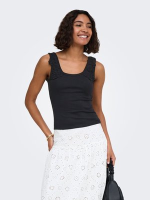 Femme souriante aux cheveux courts bouclés, portant un haut noir sans manches avec des bretelles en dentelle fleurie et une jupe blanche à oeillets, tenant un sac à main noir.