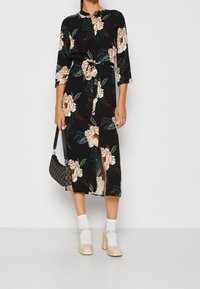 Kwiatowa sukienka midi na czarnym tle, z kremowymi i zielonymi kwiatami, z przodem zapinanym na guziki, paskiem w talii, rękawami o długości trzy czwarte oraz głębokimi bocznymi rozcięciami.