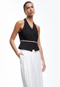 Schwarzes, gestreiftes ärmelloses Top mit V-Ausschnitt und geformtem Saum, kombiniert mit weißen, hochtaillierten Hosen; beinhaltet einen hellen Gürtel.