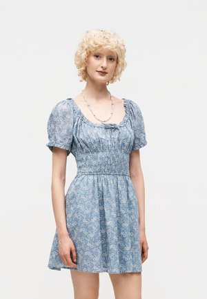 Γυναίκα με κοντά σγουρά ξανθά μαλλιά που φοράει μπλε κοντό φόρεμα με λουλούδια και φουσκωτά μανίκια, καθώς και καρφωτό κολιέ, στέκεται μπροστά σε απλό φόντο.