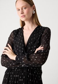Abito nero con maniche lunghe e trasparenti. Presenta un motivo a pallini argentati e un design plissettato. Scollo a V profondo e dettagli con bottoni ai polsini.
