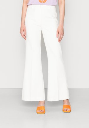 Calças brancas largas com uma textura suave, apresentando passantes para cinto e bolsos laterais. Combinadas com sapatos de salto abertos laranja para contraste.