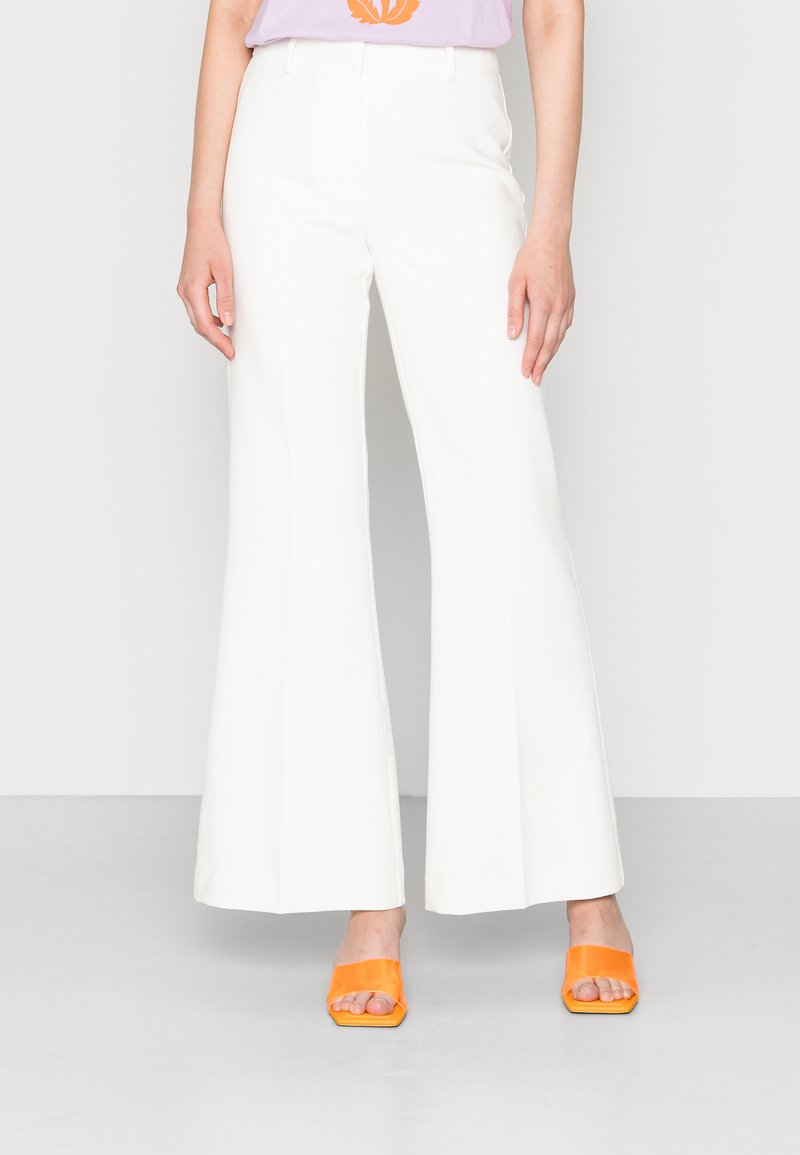 Calças brancas largas com uma textura suave, apresentando passantes para cinto e bolsos laterais. Combinadas com sapatos de salto abertos laranja para contraste.