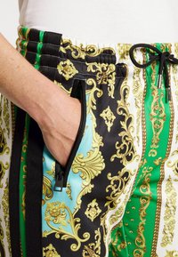 Pantalones jogger con cintura elástica en negro, blanco y verde con un patrón dorado ornamentado. Presentan bolsillos laterales con cremallera y cierre con cordón.