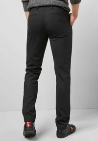 Zwarte slim-fit broek met een gladde textuur, voorzien van achterzakken en een eenvoudige tailleband, in combinatie met zwarte sneakers met rode accenten.