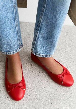 Red ballet flats made of smooth material, featuring a rounded toe and a small bow accent with gold-tone hardware. Worn with light blue jeans.