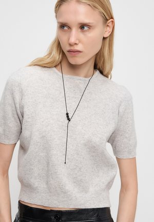 Woman with long blonde hair wearing a light grey short-sleeve sweater, black leather pants, and a black beaded necklace on a string.