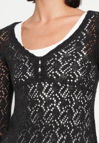 Μαύρο πουλόβερ από δαντελωτό πλεκτό ύφασμα με V-λαιμόκοψη, μακριά μανίκια και τρία διακοσμητικά κουμπιά στο μπροστινό μέρος, φορεμένο πάνω από λευκό βασικό πουκάμισο.