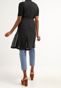 Frau in ärmelloser schwarzer Tunika über schwarzem Kurzarmoberteil, mit hellblauen Cropped-Jeans und braunen Plateausandalen, die sich wegdreht.